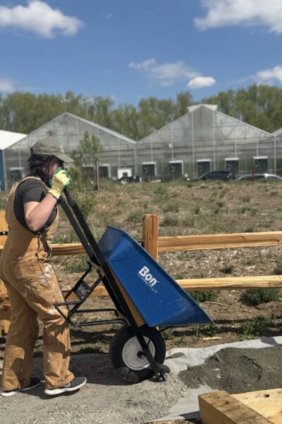 Construction worker in tan overalls and gloves operating a blue concrete mixer on a sunny outdoor construction site with a wooden barrier nearby.