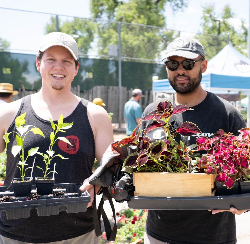 Two people at an outdoor market display plants, holding trays of young seedlings and a wooden box of colorful foliage plants.