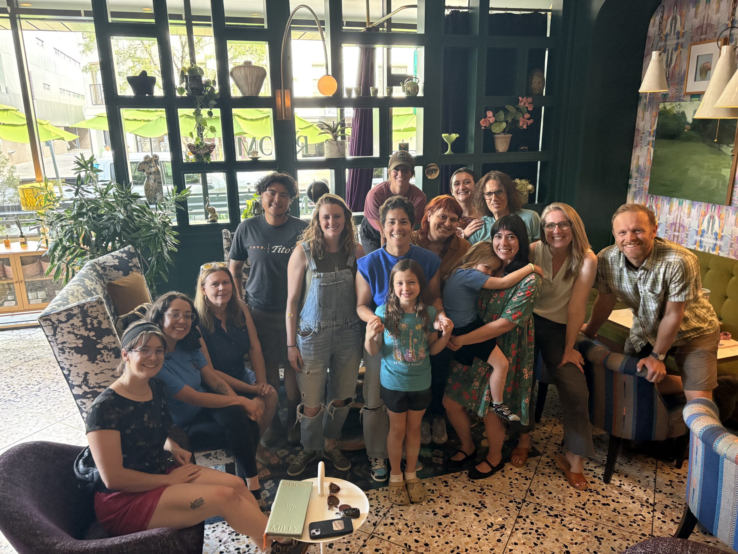 Group of smiling adults and children posing together in a cozy cafe with plants and large windows in the background