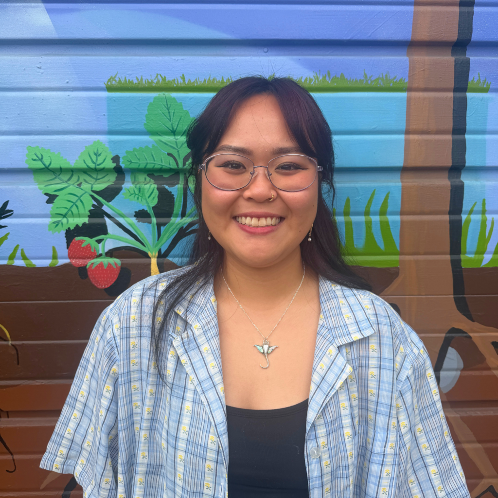 Smiling woman with round glasses in front of a colorful mural with plants and strawberries.