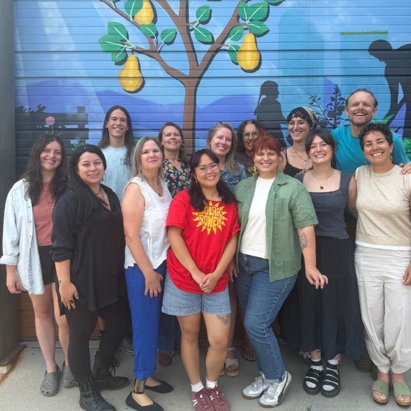 Group of around 14 adults posing and smiling in front of a bright mural with a large lemon tree image behind them (outdoor group photo).])
