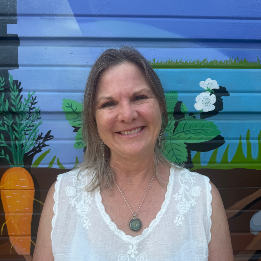 Smiling woman with light hair in a white lace top and pendant, standing before a blue mural with plants and a pumpkin.