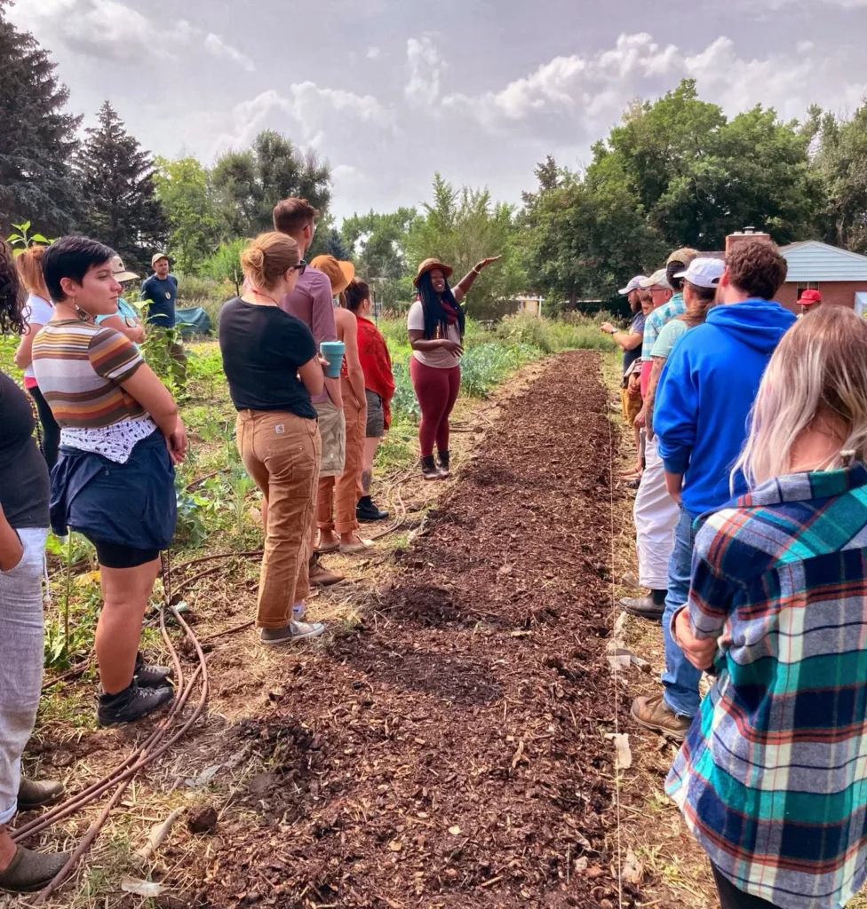 IMG-9696 - Denver Urban Gardens A woman wearing a sun hat leads a gardening demonstration to a crowd in a community garden between vegetable beds and a dirt path.