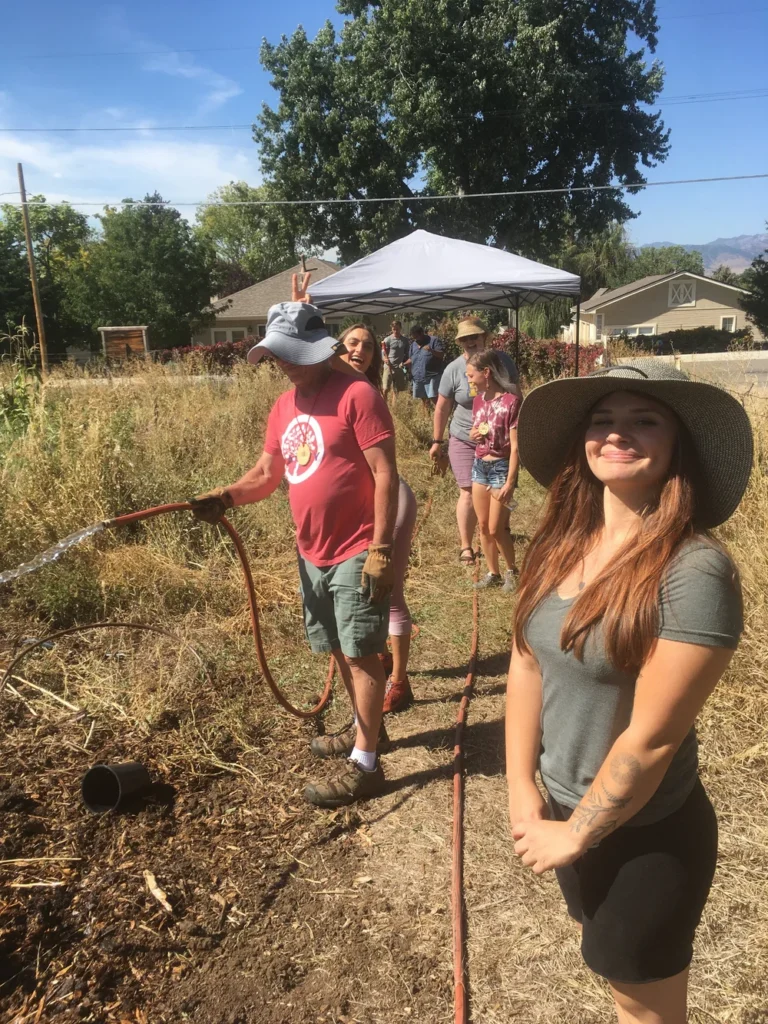 IMG_0023 - Denver Urban Gardens Group of volunteers watering a garden on a sunny day; woman in a wide-brim hat smiles in the foreground while others stand by under a canopy in the background.
