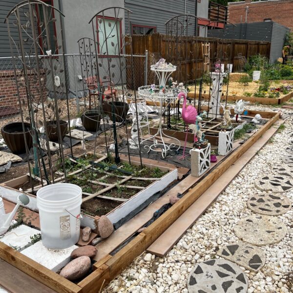 Garden with decorative metal arch trellises over raised wooden planter beds and scattered garden statues and a pink flamingo ornament in a gravel yard.