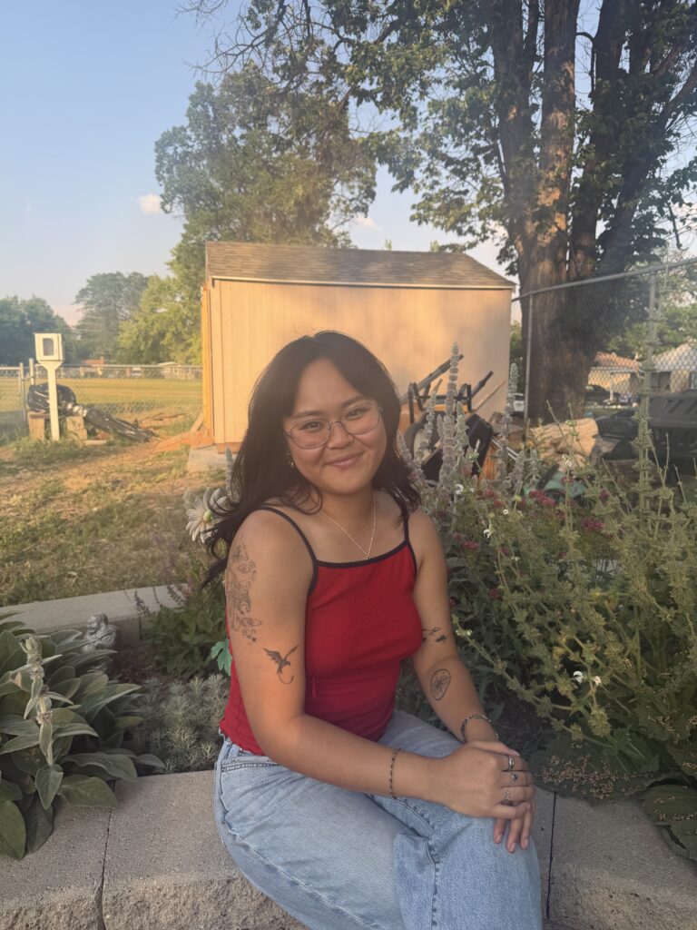 Smiling woman with glasses wearing a red sleeveless top sits on a garden wall, displaying tattoos on her arms with greenery and a shed behind her.