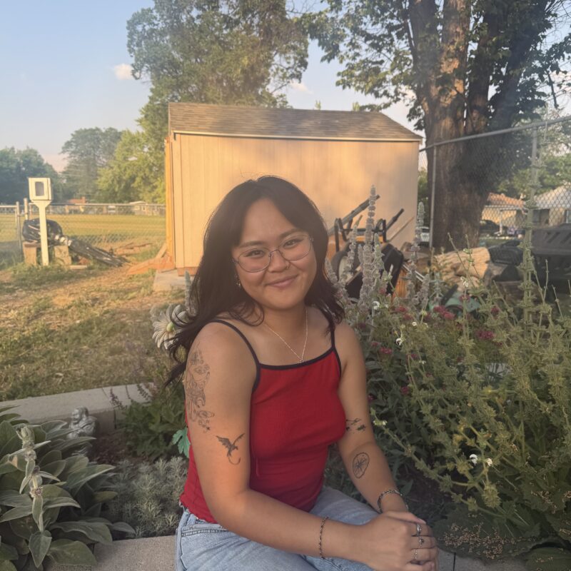 Smiling woman with glasses wearing a red sleeveless top sits on a garden wall, displaying tattoos on her arms with greenery and a shed behind her.