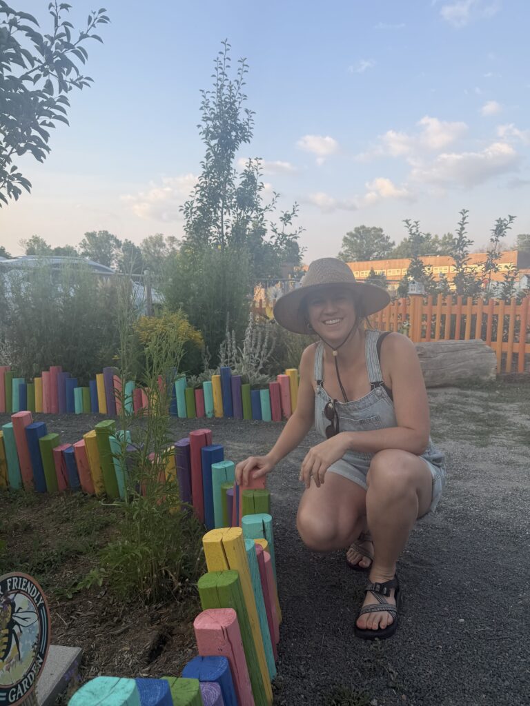 Smiling woman wearing a straw hat and denim overalls crouches beside a rainbow-painted wooden fence in a garden.