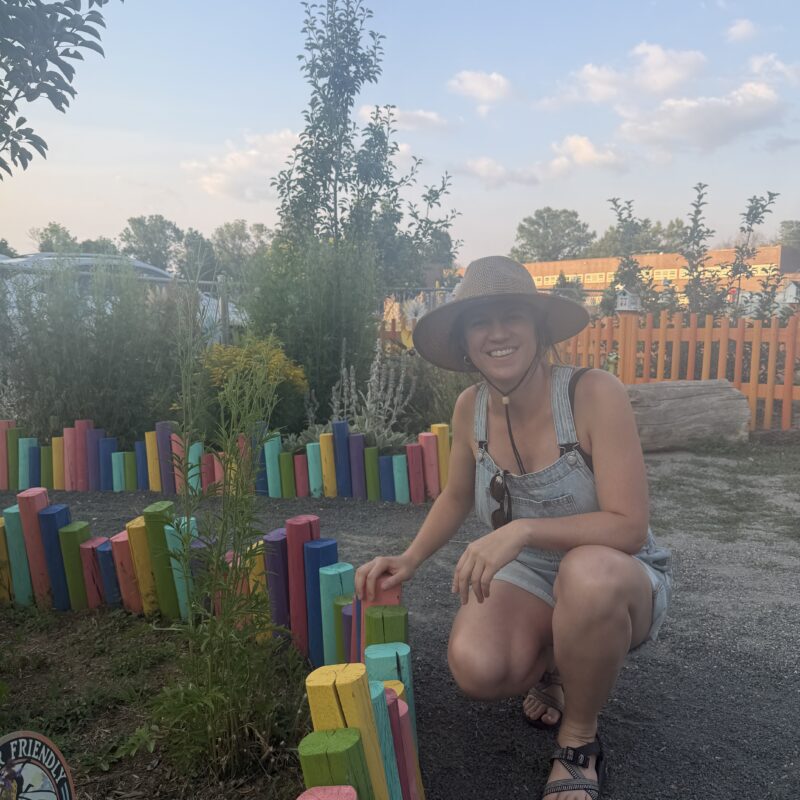 Smiling woman wearing a straw hat and denim overalls crouches beside a rainbow-painted wooden fence in a garden.
