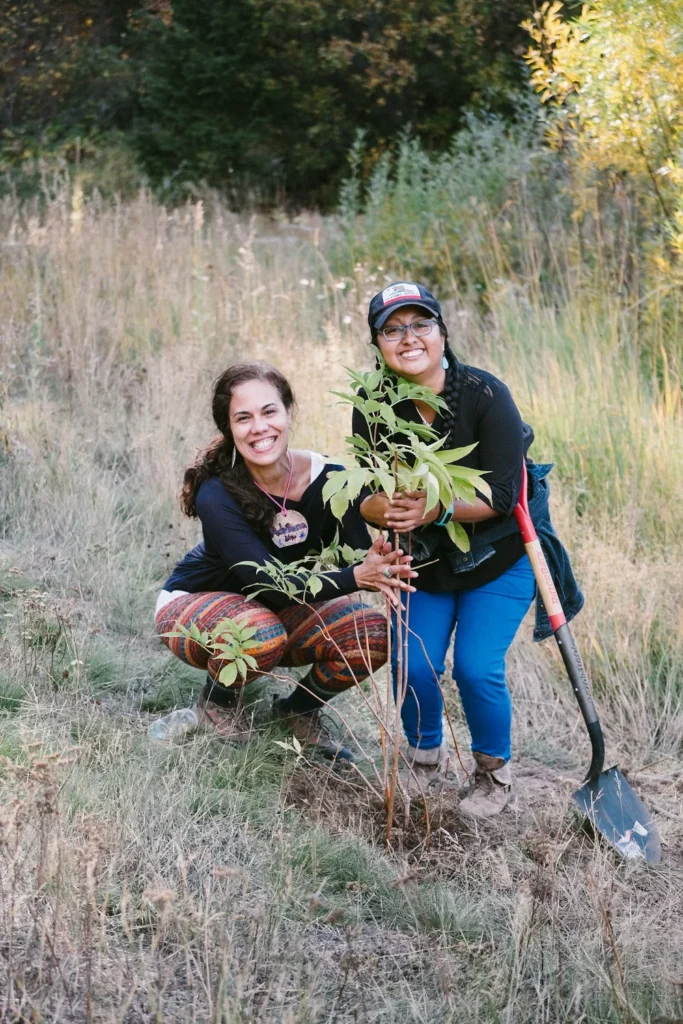 Perma-92 - Denver Urban Gardens Two smiling women plant a young tree together in a grassy field, one holding the sapling while a shovel rests nearby peaceably behind them