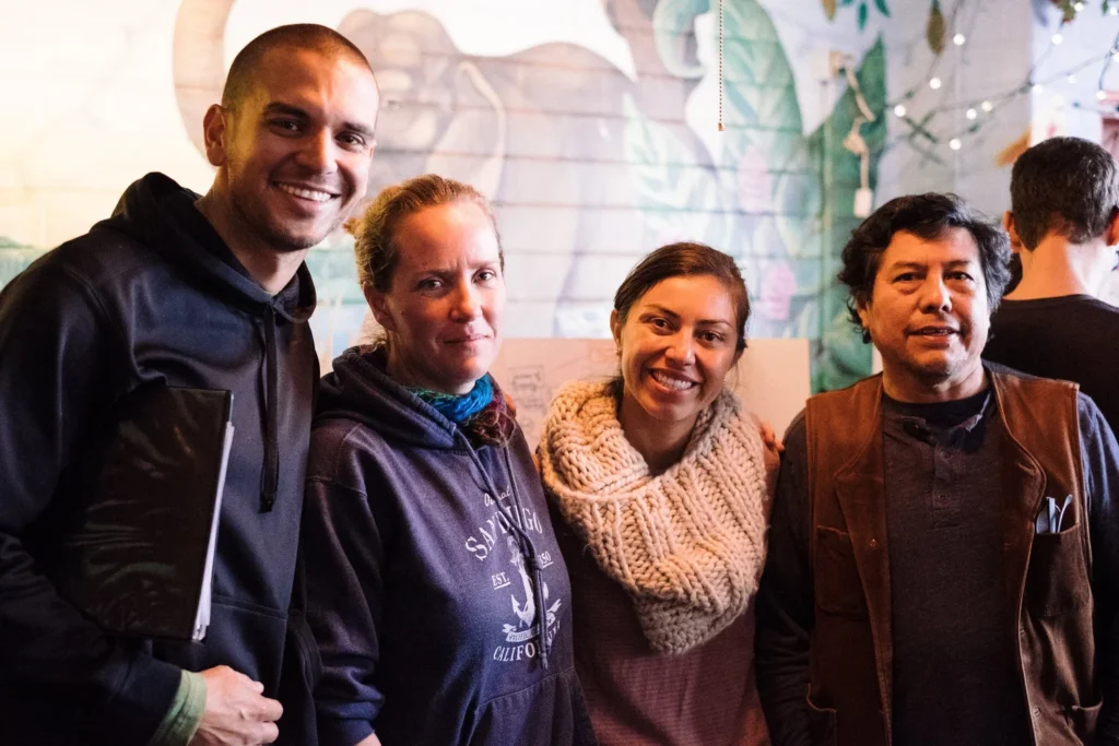 Permaculture-51+1 - Denver Urban Gardens Four people posing together indoors, smiling at the camera with a colorful mural behind them, left person holding a folder/ notebook.