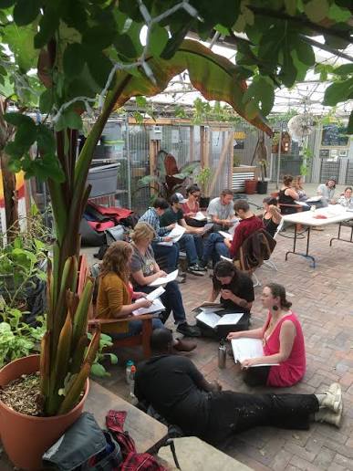 images 2 - Denver Urban Gardens Group of adults seated in a greenhouse courtyard, reading papers and discussing together in a plant-filled setting
