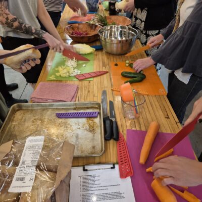 Group of people chopping vegetables around a wooden table on cutting boards, with cucumbers, carrots, bowls, and a recipe sheet visible.