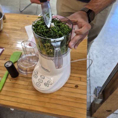 Person with a wristwatch holds a knife over a white blender, chopping green herbs inside the blender's jar on a wooden table
