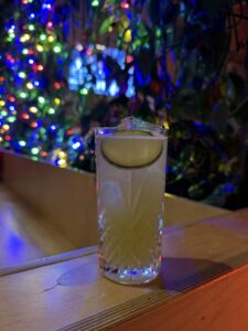 Tall, textured glass with ice and a lime wedge on a wooden bar, colorful bokeh lights in the background (bar scene).