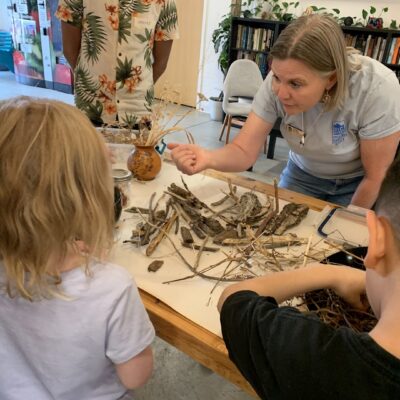 Instructor guides children as they arrange sticks and wood pieces on a table during a hands-on craft in a classroom/library setting.