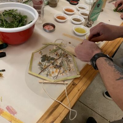 Person making a small pressed-flower art piece at a craft table, with a hexagonal frame, dried flowers, and bowls of colored paints nearby.