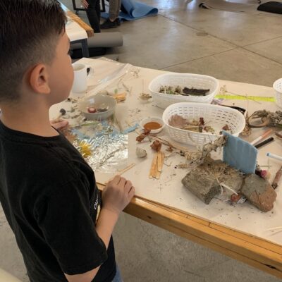 Young child standing at a table exploring a natural-material art project with shells, twigs, and stones.