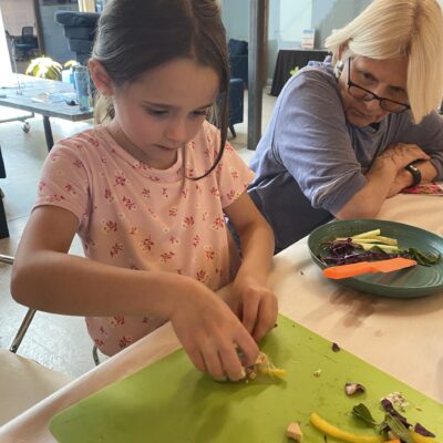 A young girl chopping vegetables on a green cutting board at a table, with an older woman nearby watching.