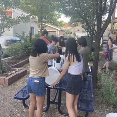 Group of young people gathered around a blue metal table outdoors, handling white buckets in a courtyard under trees.