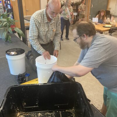 Older man with a white beard pours material from a bucket into a large black bin while a younger man assists nearby in a community workshop setting.