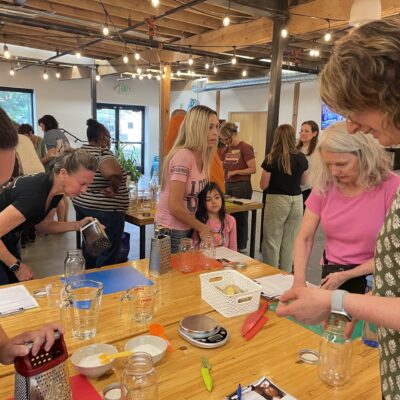 Group of people around a long wooden table in a bright workshop, preparing ingredients with jars, scales, and cutting tools nearby.