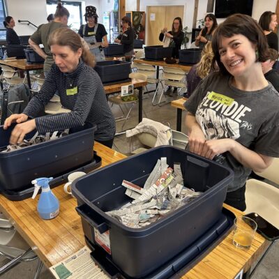 People in a workshop sorting shredded newspaper into plastic bins at long tables, wearing name tags and casual clothes.