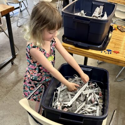 Young girl in a colorful dress sorts shredded newspaper from a bin in a workshop space with tables and chairs.
