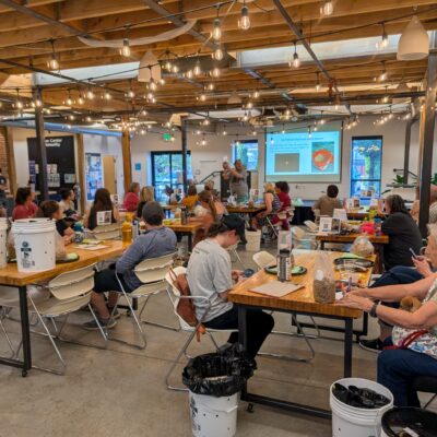 Community workshop in a bright, rustic hall with string lights; people sit at long tables listening to a presenter at the front.