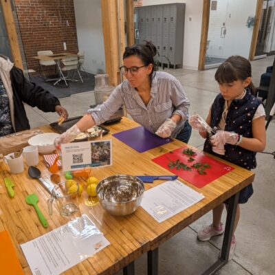Two people cooking at a wooden workshop table, chopping herbs with gloves in a modern, loft-style kitchen space.