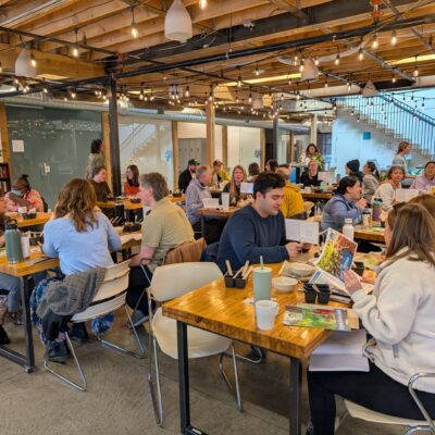 Crowded open-plan cafe or coworking space with long wooden tables, people eating and chatting under warm string lights.