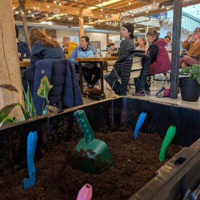 Indoor workshop space with string lights and people seated at tables; a large planter bed filled with soil and colorful plastic shovels in the foreground.