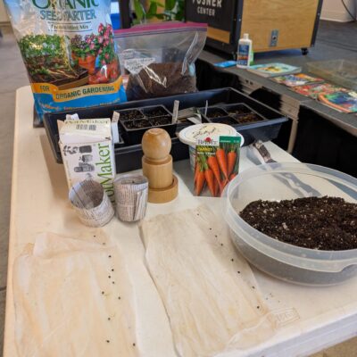 A seed-starting station with organic potting mix bag, seed trays, and a large tub of soil on a table at a workshop or classroom.