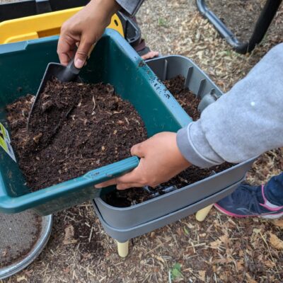 Person uses a small shovel to add soil to a green planter outdoors.