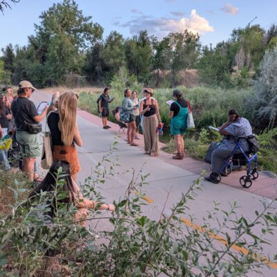 Group of people gathered along a paved trail beside a small waterway, some holding papers or booklets.