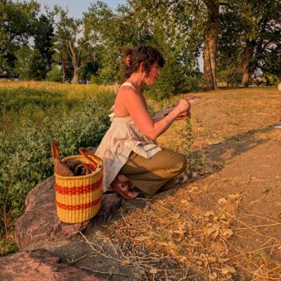 Woman with dark curly hair tied back sits on a large rock, hands busy with a small object, a woven basket beside her, in a sunlit park with trees in the background.