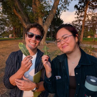 Two friends smiling at the camera in a park, holding green leaves, with trees and a sunset sky behind them.