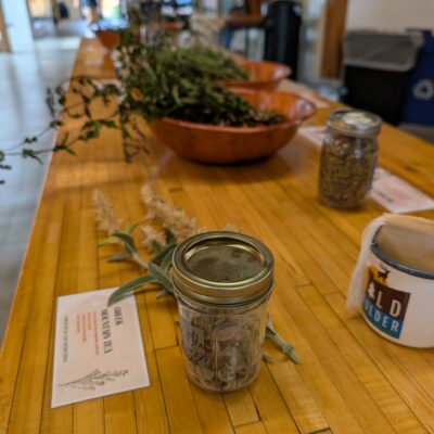 Blurred indoor workspace showing a long wooden table with glass jars of dried herbs and greenery; a person moves in the background, suggesting an herb workshop.