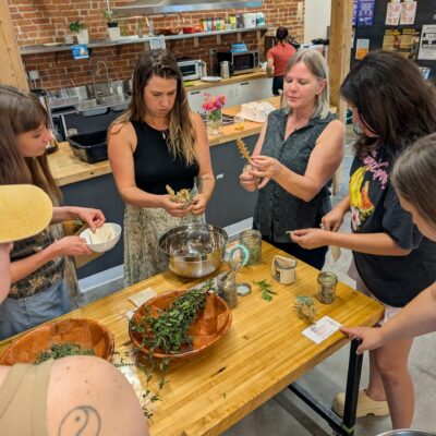 People gathered around a wooden worktable, shredding herbs in a kitchen workshop with brick walls and flags overhead.