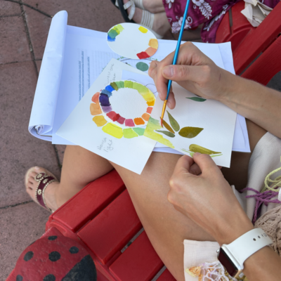 Person paints a rainbow color wheel and leaf designs on white cards with a blue-handled brush, outdoors on a red bench.
