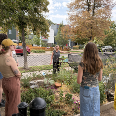 A woman in a sleeveless top guides a small group in a community garden, gesturing toward the plants and raised beds as neighbors listen nearby with a fence and houses in the background.