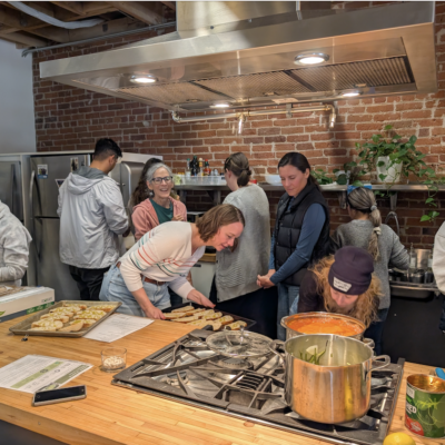 Several people in a kitchen class work around a large stainless-steel stove, preparing trays of pastries on a wooden island.