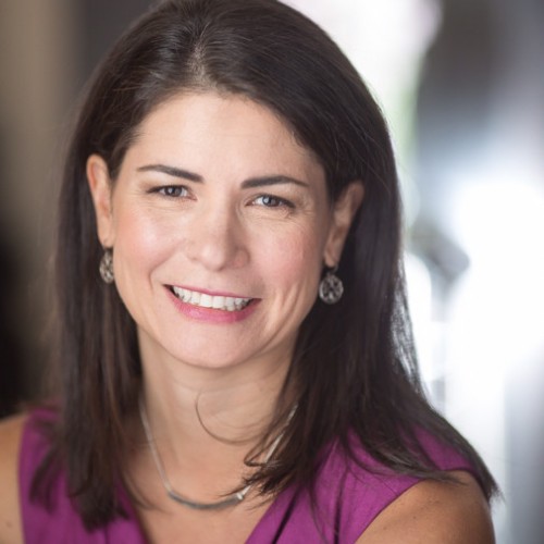 Portrait of a smiling woman with shoulder-length brown hair, wearing a purple sleeveless top and silver earrings.