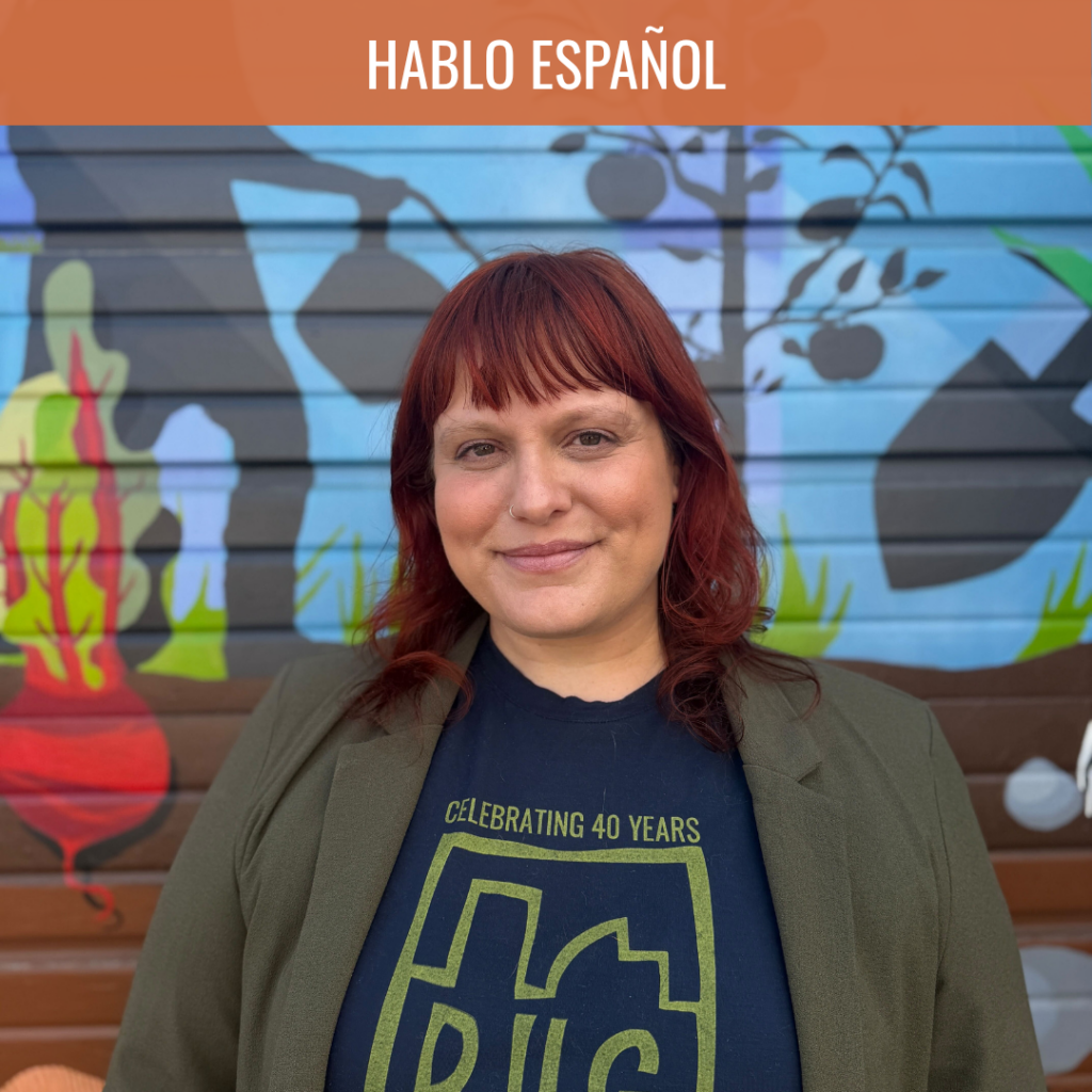 Portrait of a woman with red hair and a nose ring, standing in front of a colorful graffiti mural, wearing a navy shirt and olive blazer.