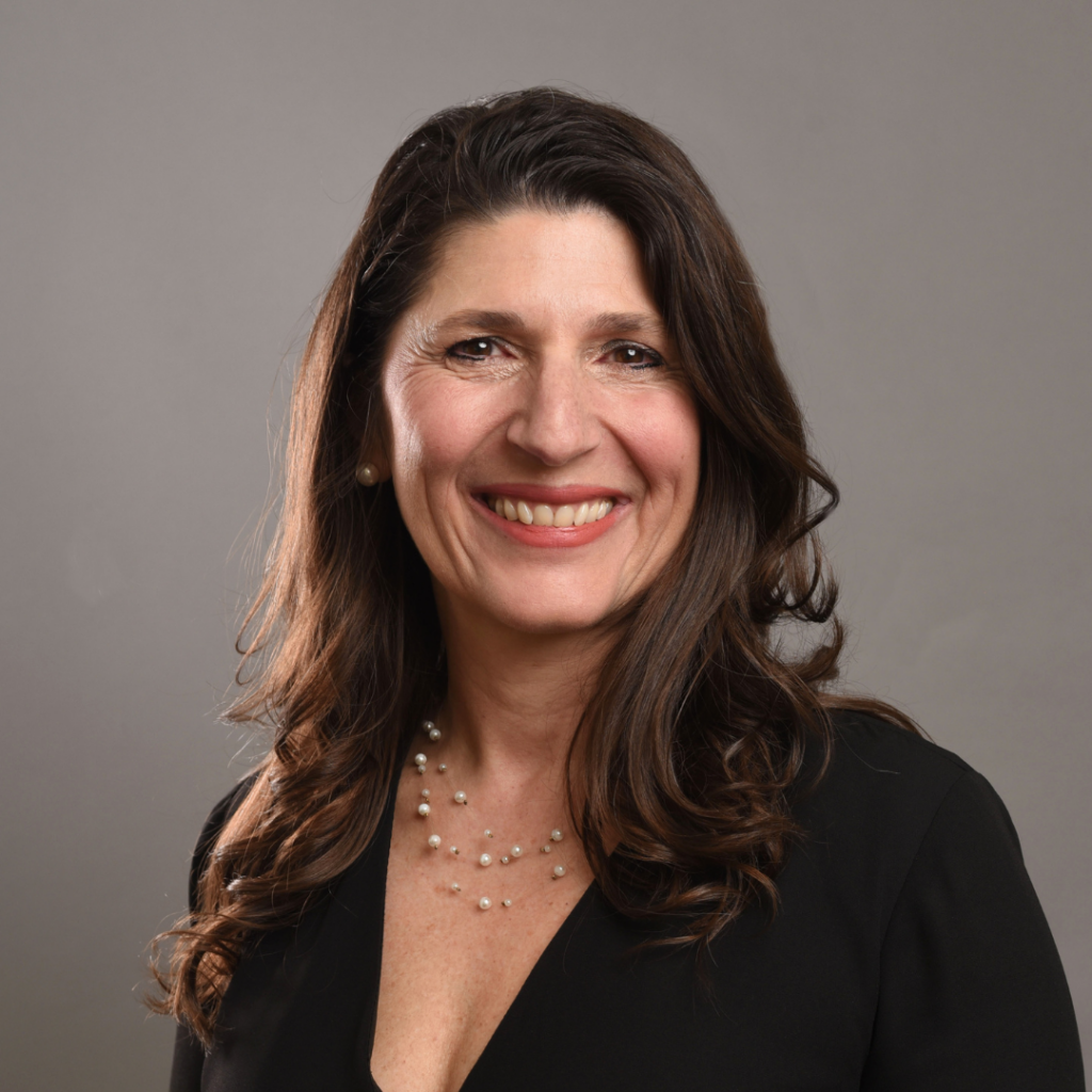 Smiling woman with dark hair in a black blouse, wearing a pearl necklace, against a neutral gray backdrop.