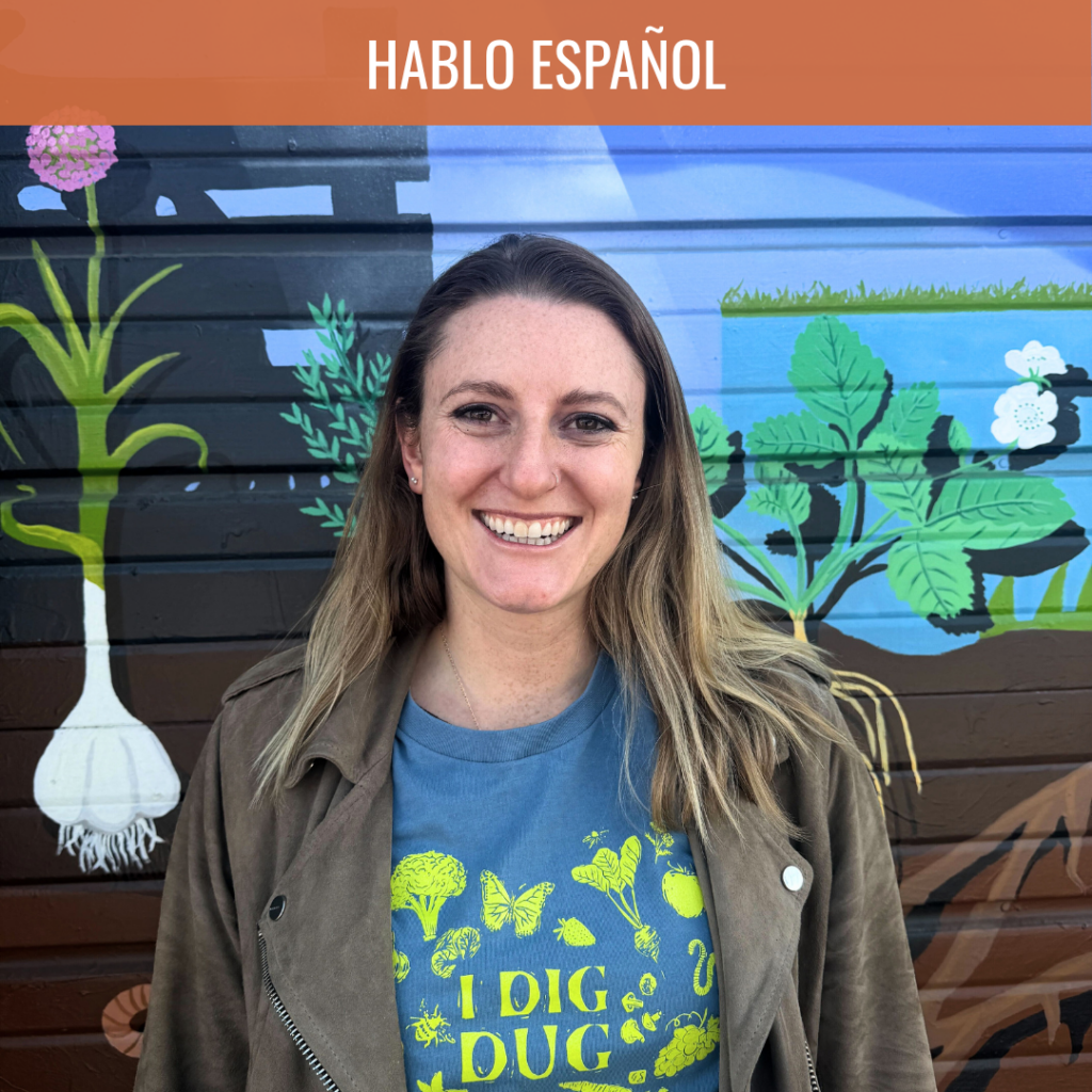 Smiling woman in a teal shirt and brown jacket stands before a colorful mural, suggesting a Spanish-language program.