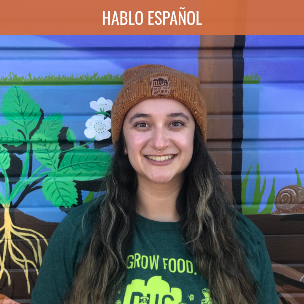 Smiling young woman in a brown knit beanie with nose ring, standing in front of a colorful mural garden background.